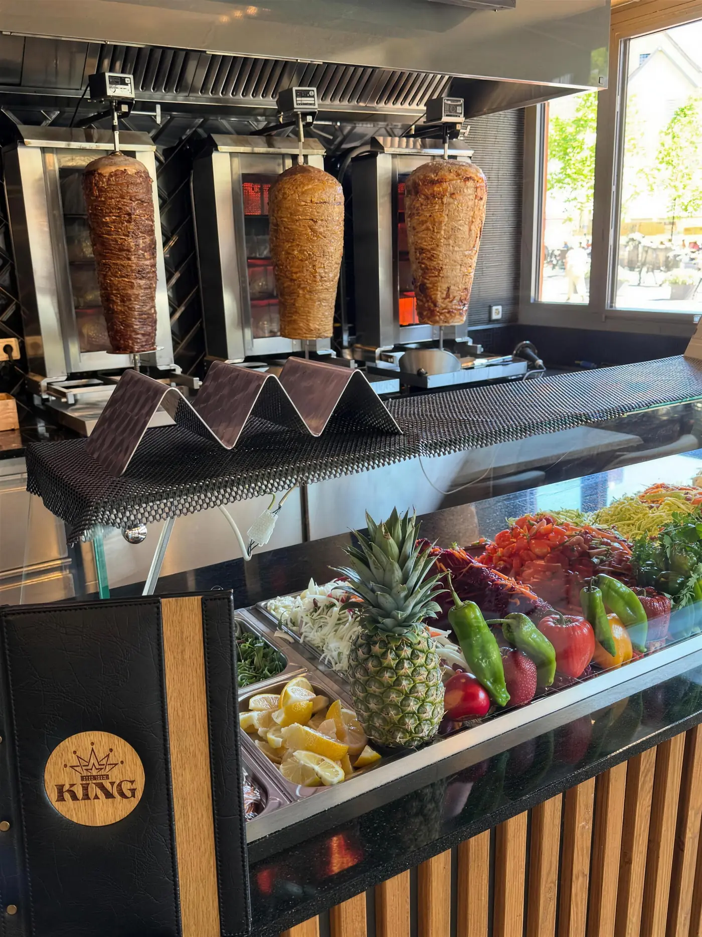 Theke im King Kebap Lünen mit drei Döner-Spießen am Grill und frischem Gemüse, Zitronen und Ananas in der Auslage